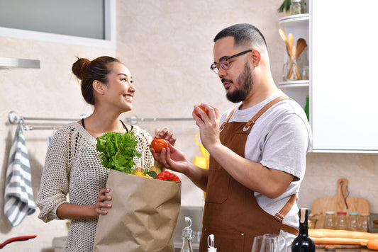 Loving young Asian couple cooking in kitchen making healthy food and holding grocery shopping bag with vegetables together feeling fun