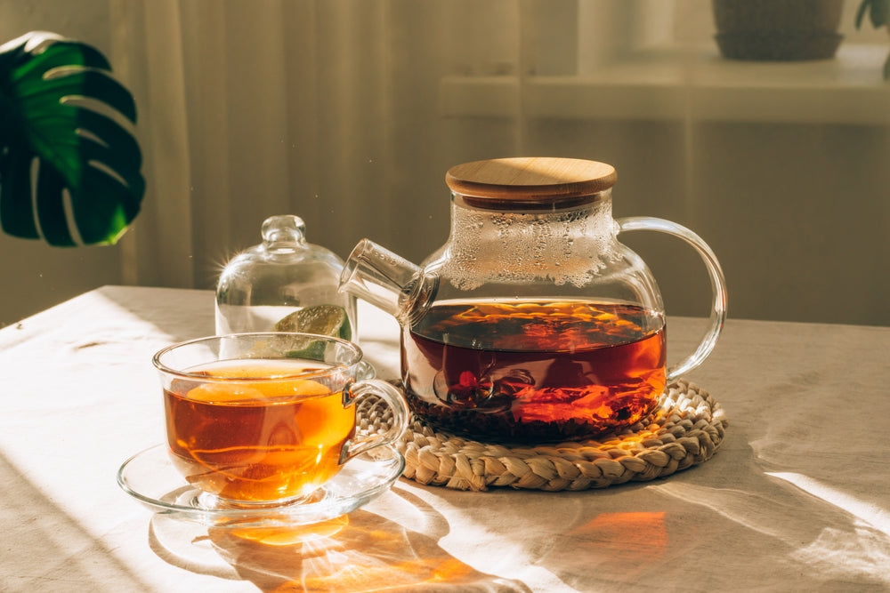 Glass teapot with brewed tea, cup of tea and lime under warm sunlight on the table with green leaf in the background